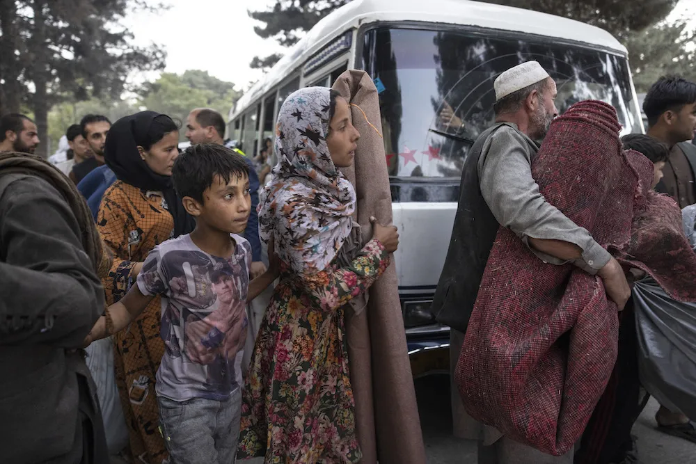 A group of people carry rugs, fabrics, and other items as they walk past a white van.
