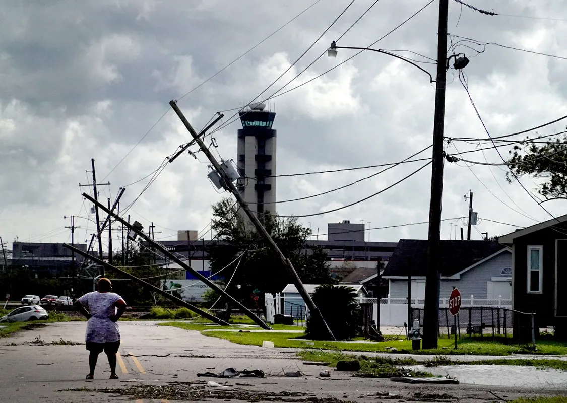 Una mujer está parada en una calle de Luisiana que ha sido dañada por el huracán Ida. Varios postes telefónicos están caídos y hay escombros en la calle.