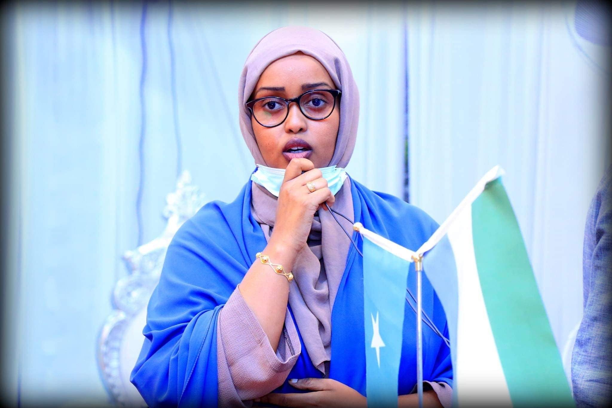 Una joven mujer somalí que lleva un velo morado, gafas negras redondas y una chaqueta azul da un discurso