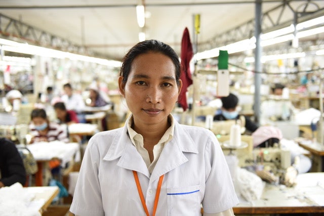 A Cambodian woman wearing a white collared shirt looks directly at the camera. She is standing in a garment factory, and behind her are women working at tables and using sewing machines.