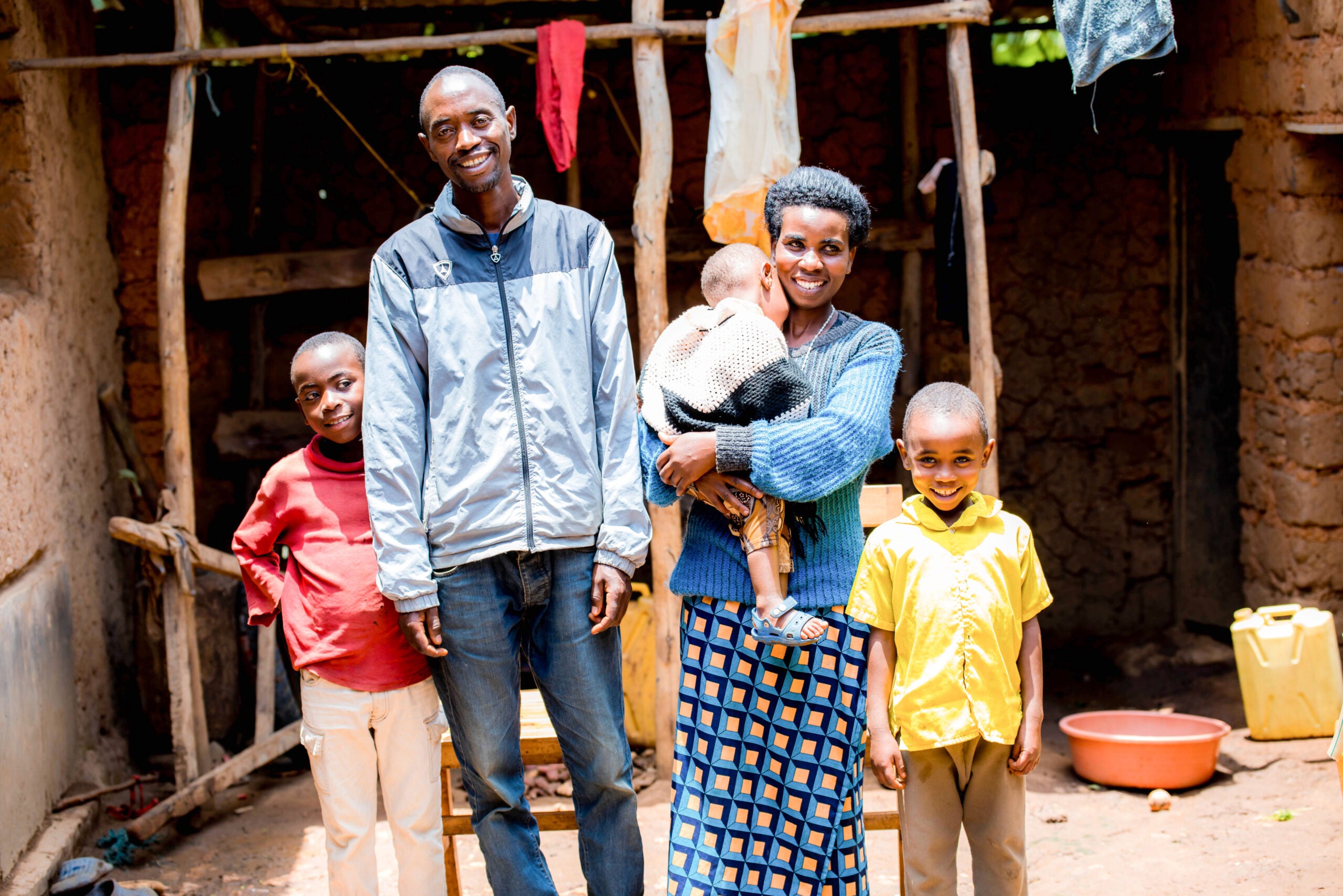 A Rwandan son (red shirt), dad (gray jacket), baby (white shirt) held by mom (blue and gray sweater), and son (yellow shirt) pose for a portrait