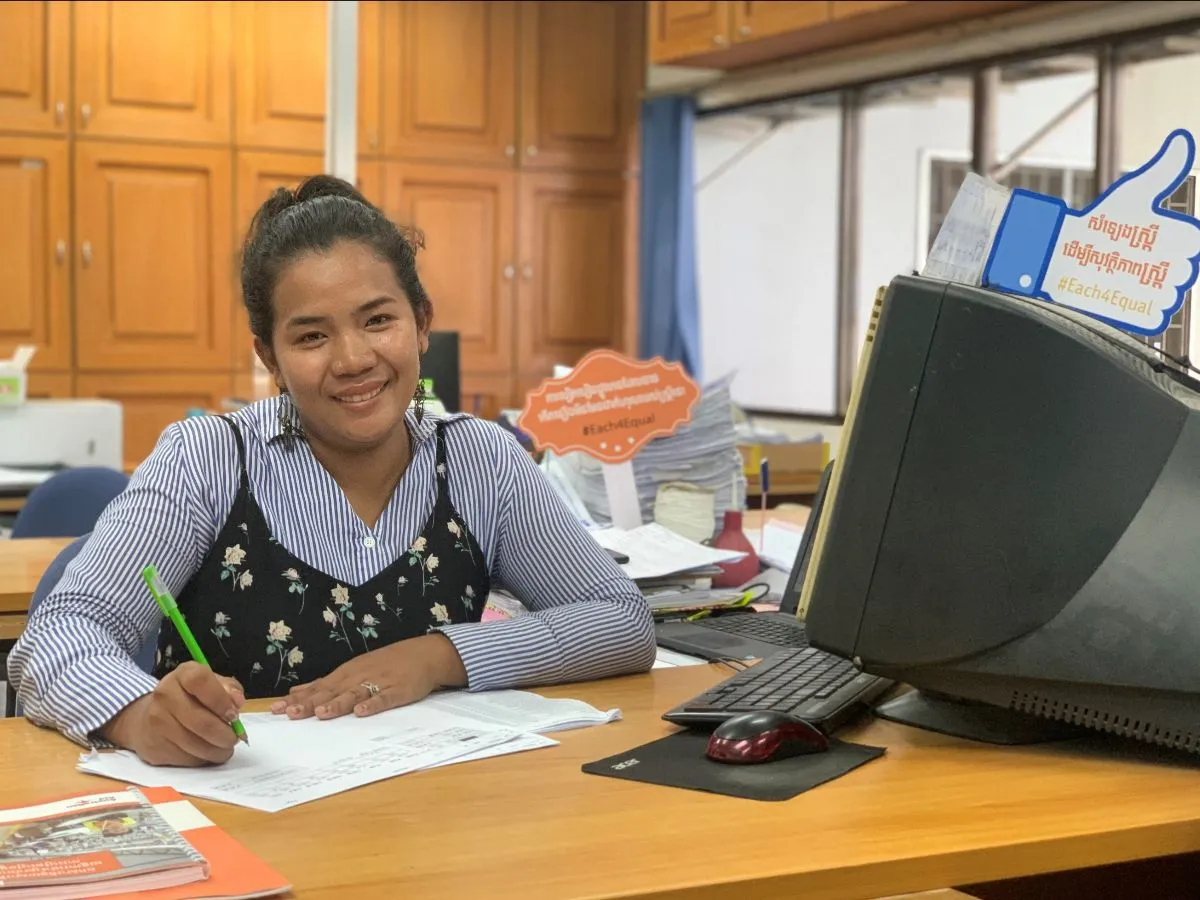 Una mujer sonríe en un escritorio mientras llena un formulario en papel. Ella está sentada en un escritorio frente a una computadora.