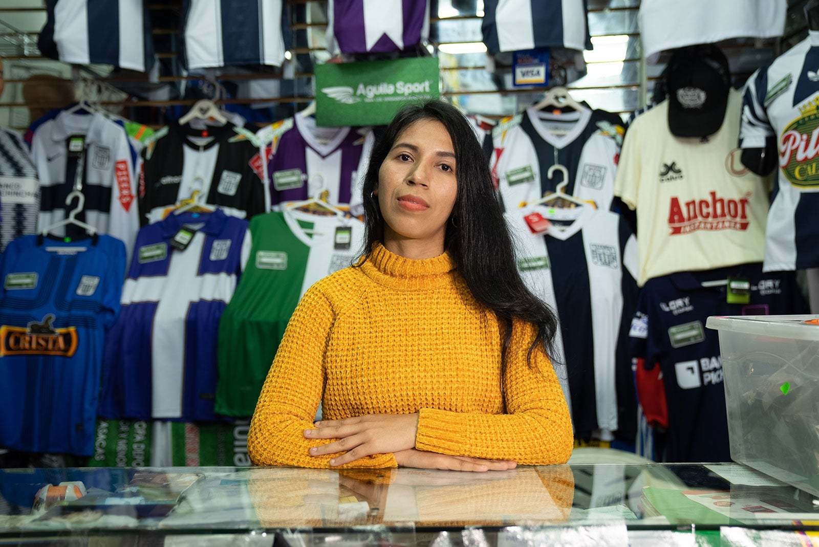A Peruvian woman wearing a gold sweater stands at the counter in her store. Behind her are rows of hanging jerseys.