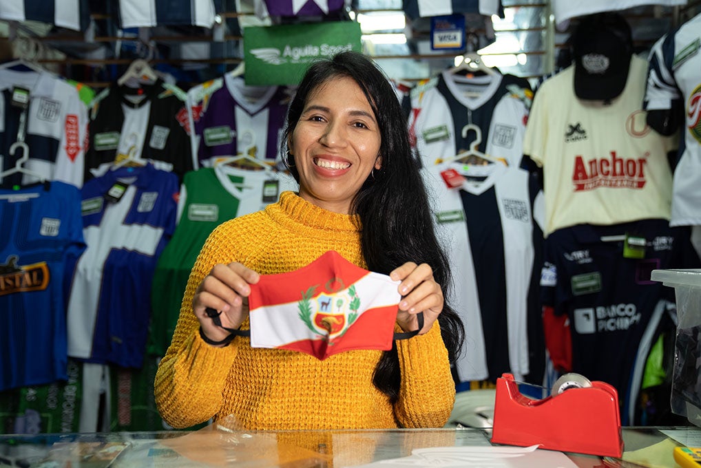 Una mujer sonriente sostiene una mascarilla roja y blanca con la bandera peruana.