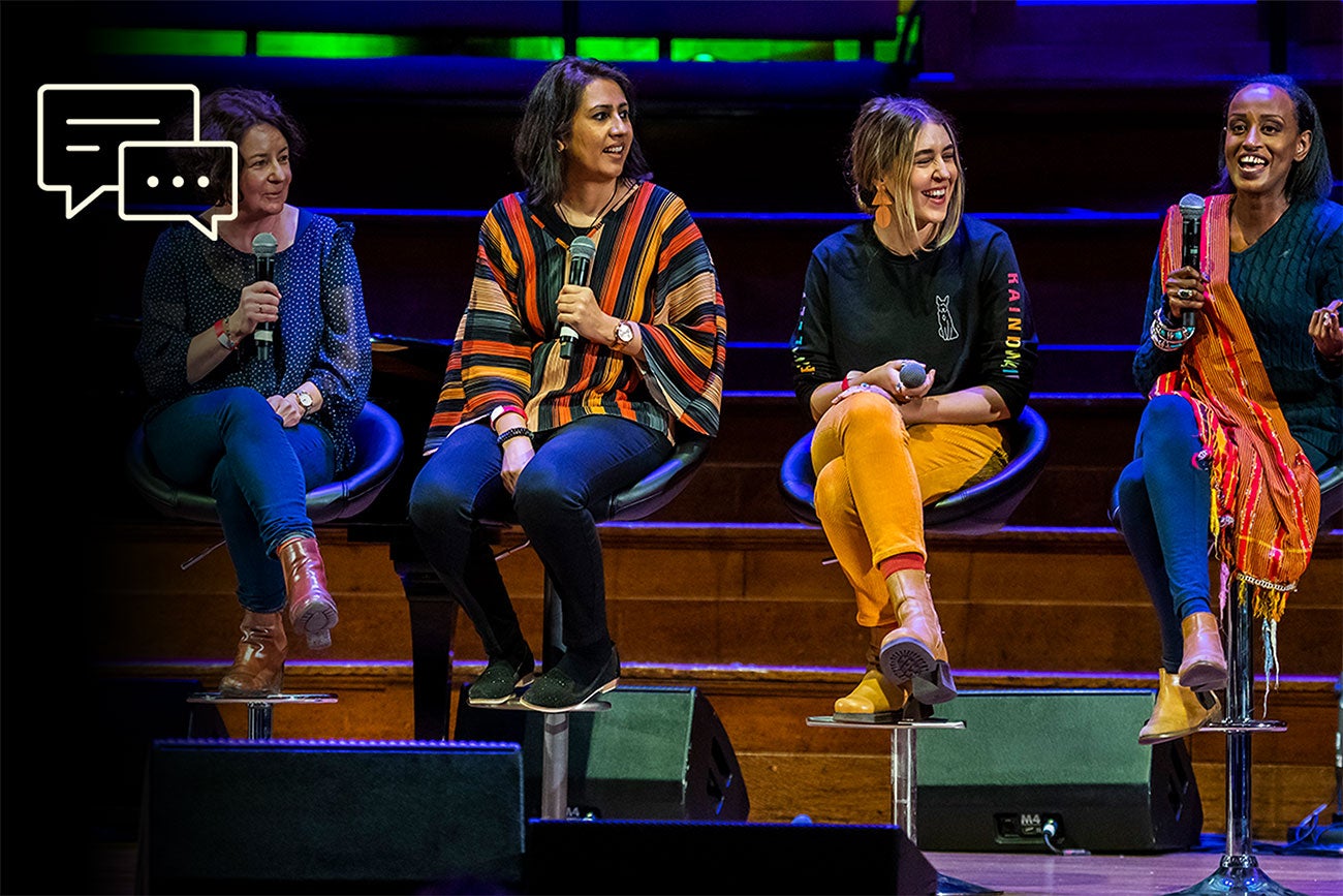 Four women participate in a panel on stage. Three are listening eagerly while a fourth speaks.