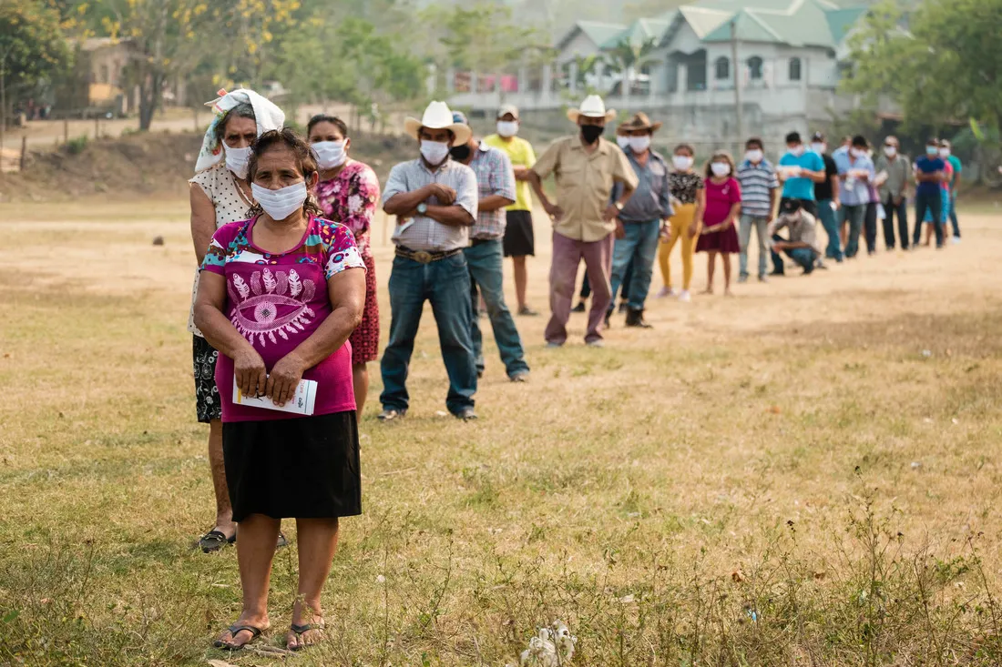 People in Villanueva, Honduras, wait in a long, single-file line in a grassy field. Each of them is wearing a white face mask.