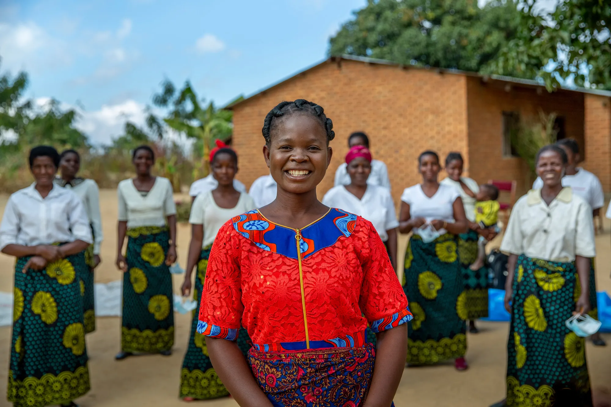 A Malawian woman wearing a bright red shirt smiles in the center of the photo. Behind her, a group of women in matching patterned skirts and white shirts stand in a semi circle.