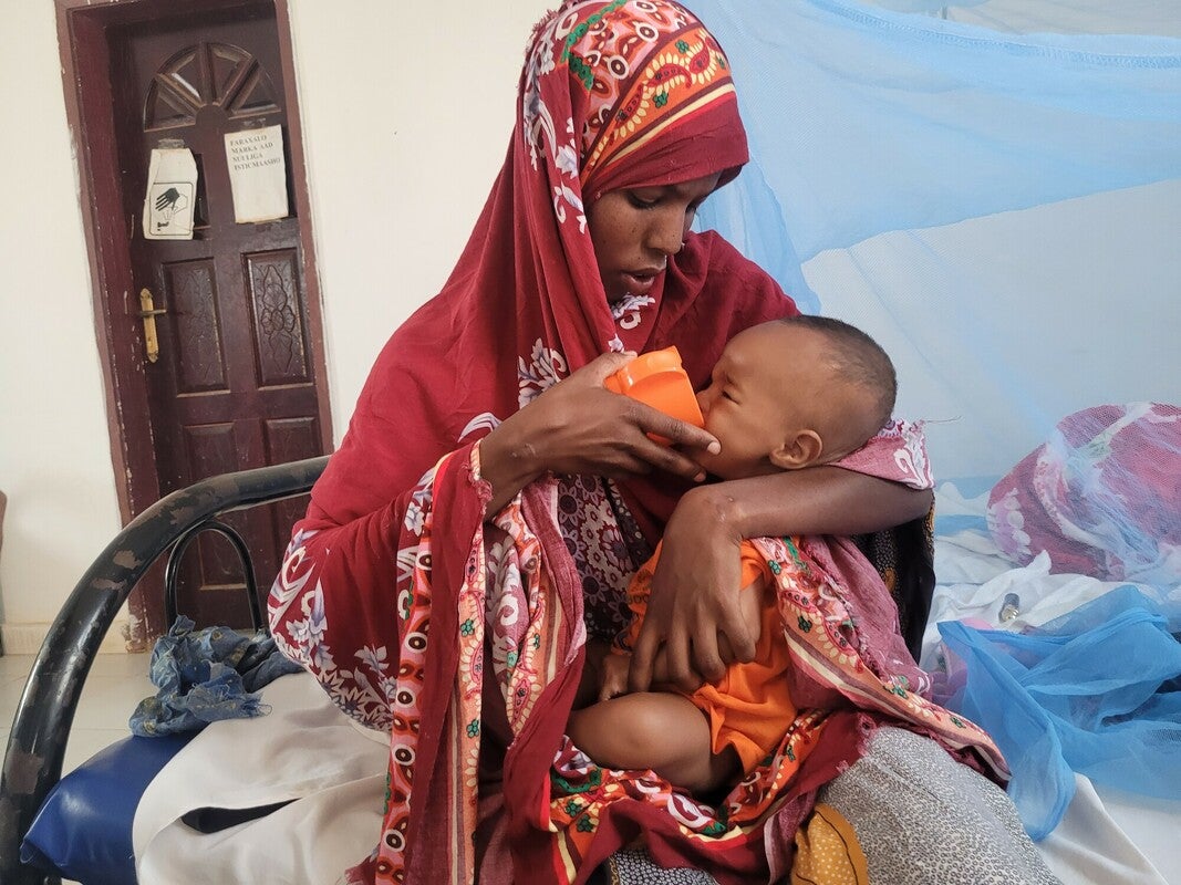 A Somali woman wearing a long burgundy head scarf holds a child in her lap and feeds them out of an orange cup.