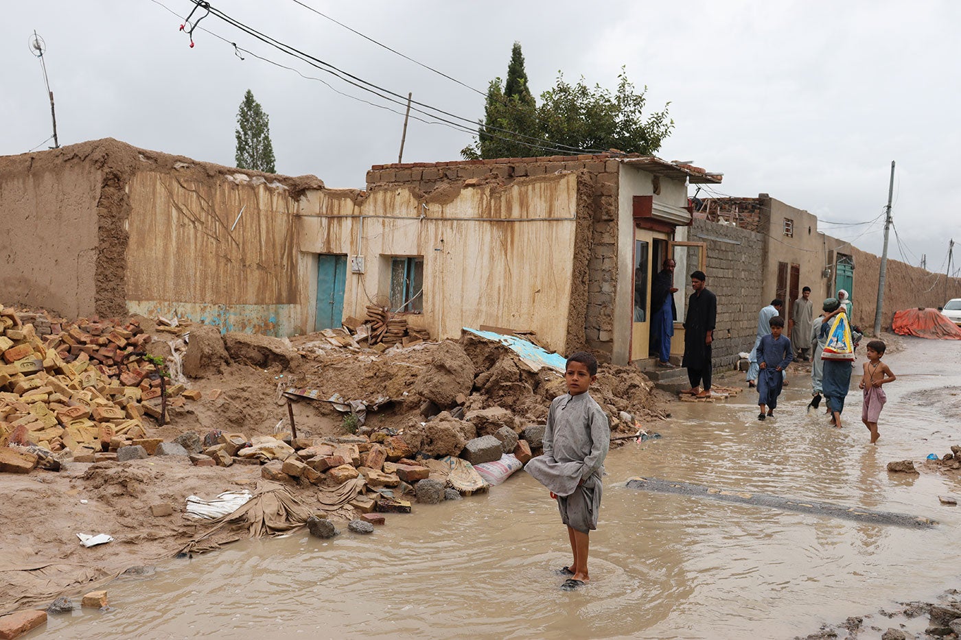 A boy walks through a flooded dirt path alongside some rubble. Behind him, a few adults and children walk together.
