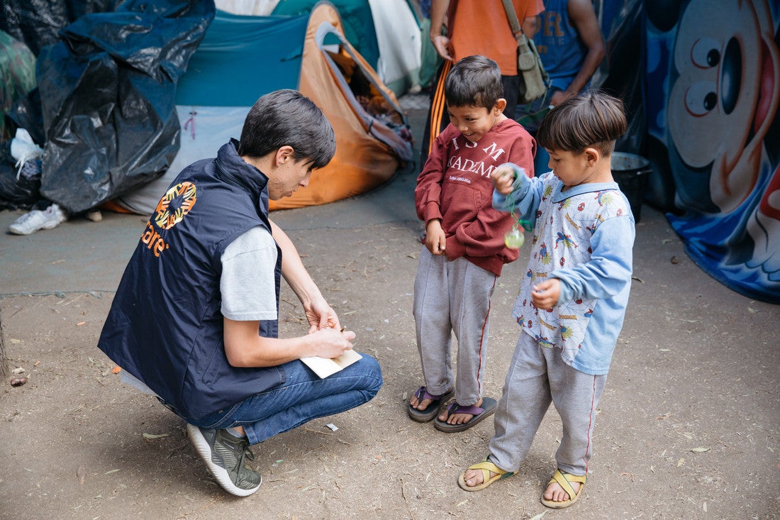 A woman wearing jeans and a navy CARE vest kneels down and writes information down on a notepad. Standing before her are two young boys. One of the boys is laughing, while the other plays with a yo-yo.