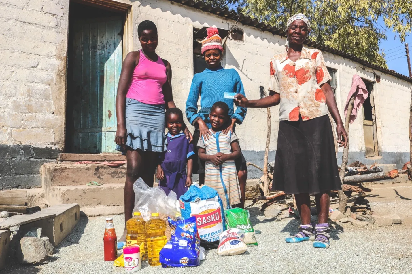 Three women and two children stand behind a bundle of food. One of the women holds up a card.