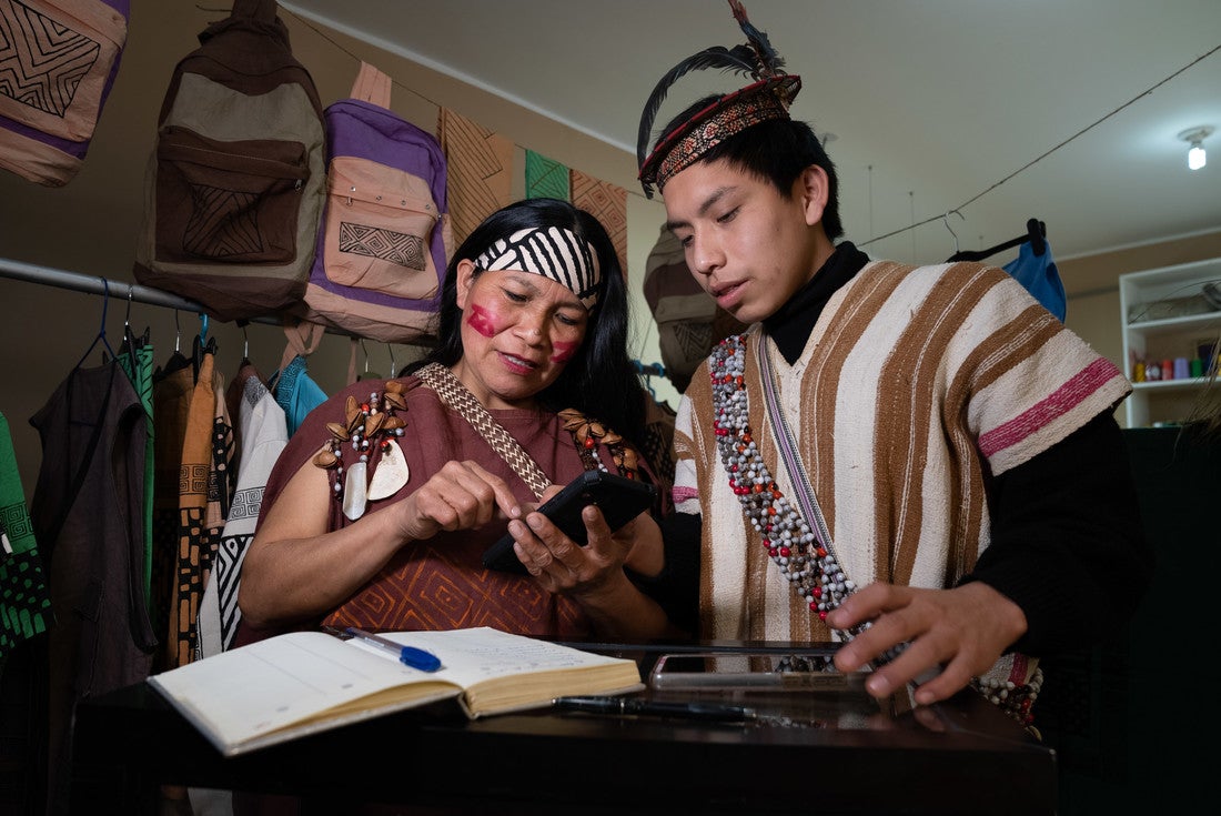 A Peruvian woman with decorative red paint streaked across her cheeks stands behind a counter with a young man. She is pressing buttons on a calculator while talking. In front of them, an open notebook lies on the counter.