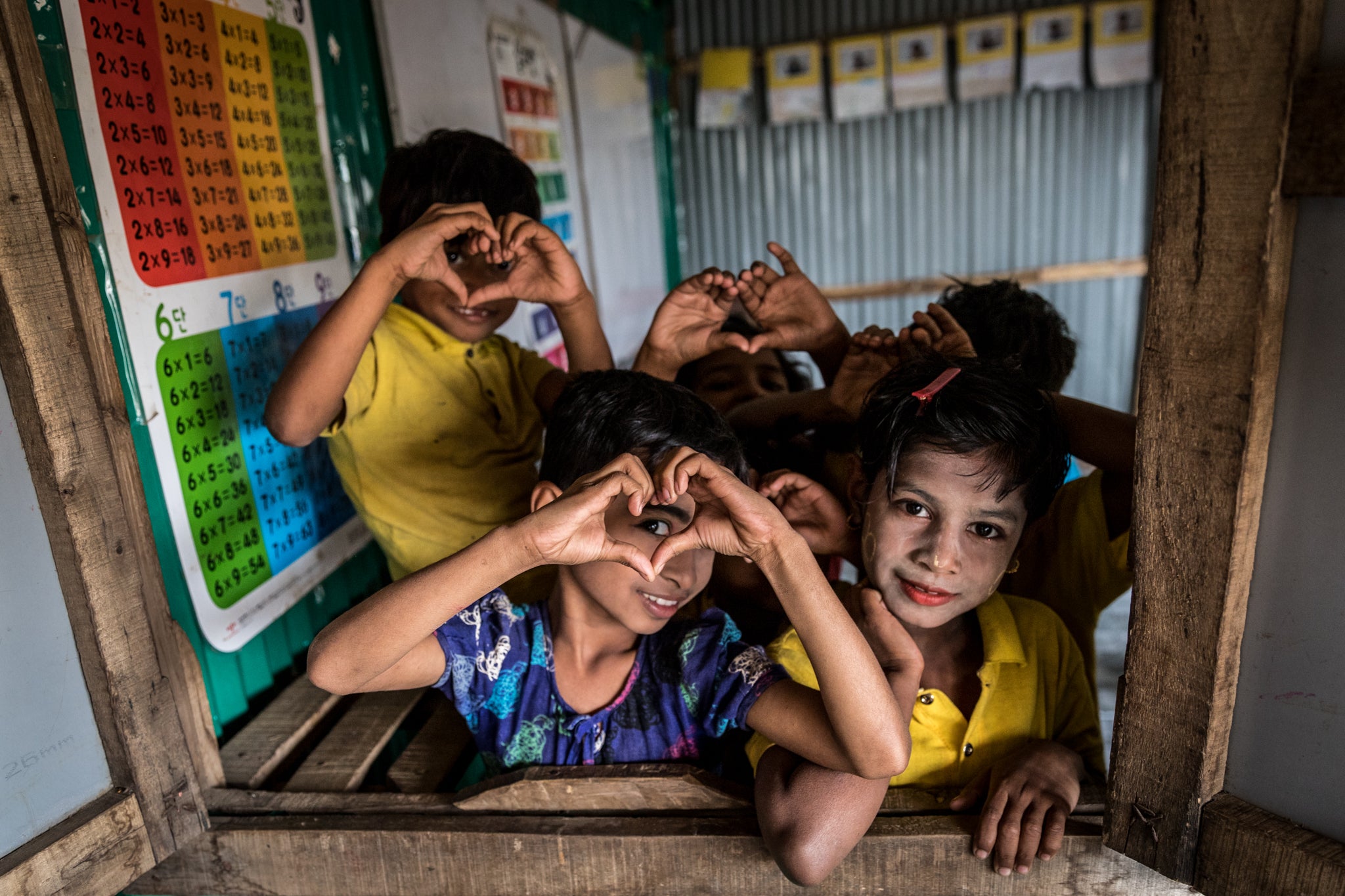 A group of kids crowd together to look out of a schoolroom window. Three of the kids are holding up hands in the shape of hearts in front of their faces.