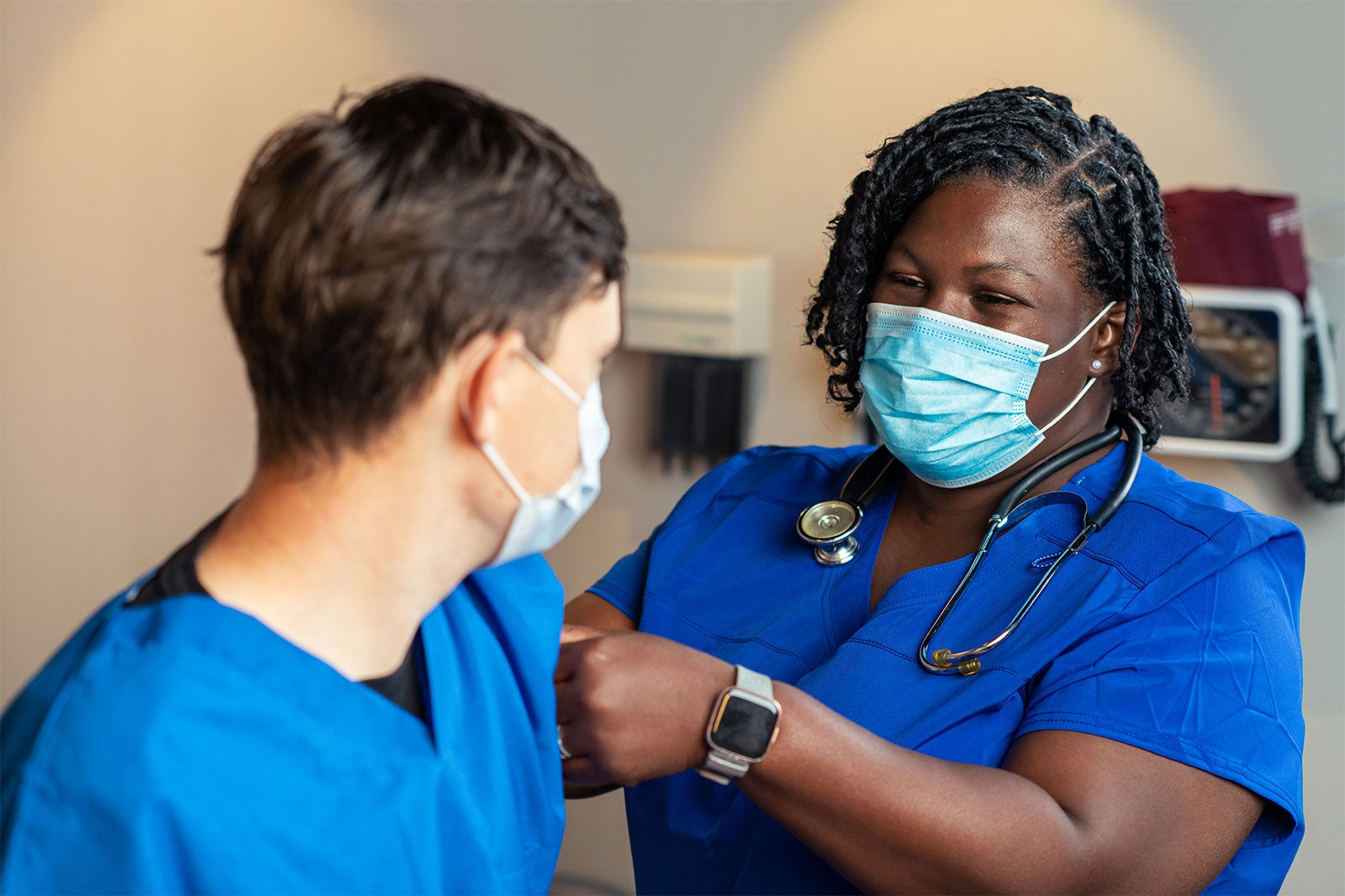 A Black female healthcare worker puts a band-aid on a young white man's arm. Both are wearing blue surgical facemasks.