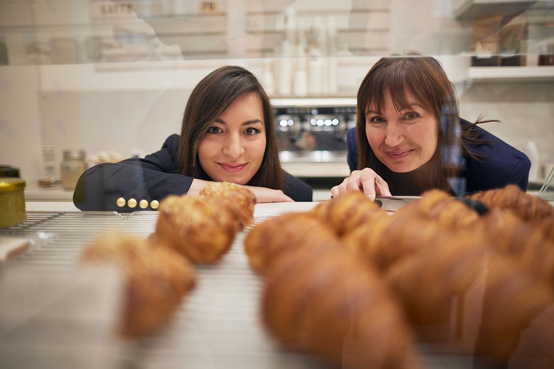 Two women peer into a display tray to view croissants and other pastries.
