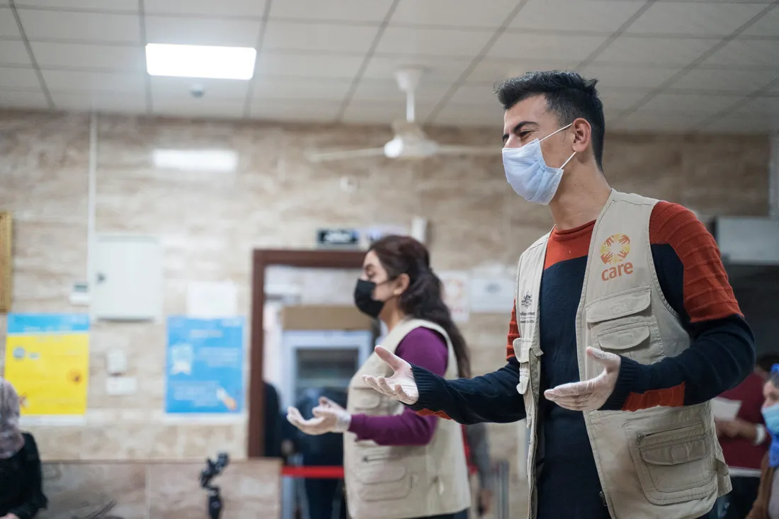 Um homem e uma mulher, cada um usando uma máscara facial e um colete cáqui CARE, demonstram práticas seguras de higiene.