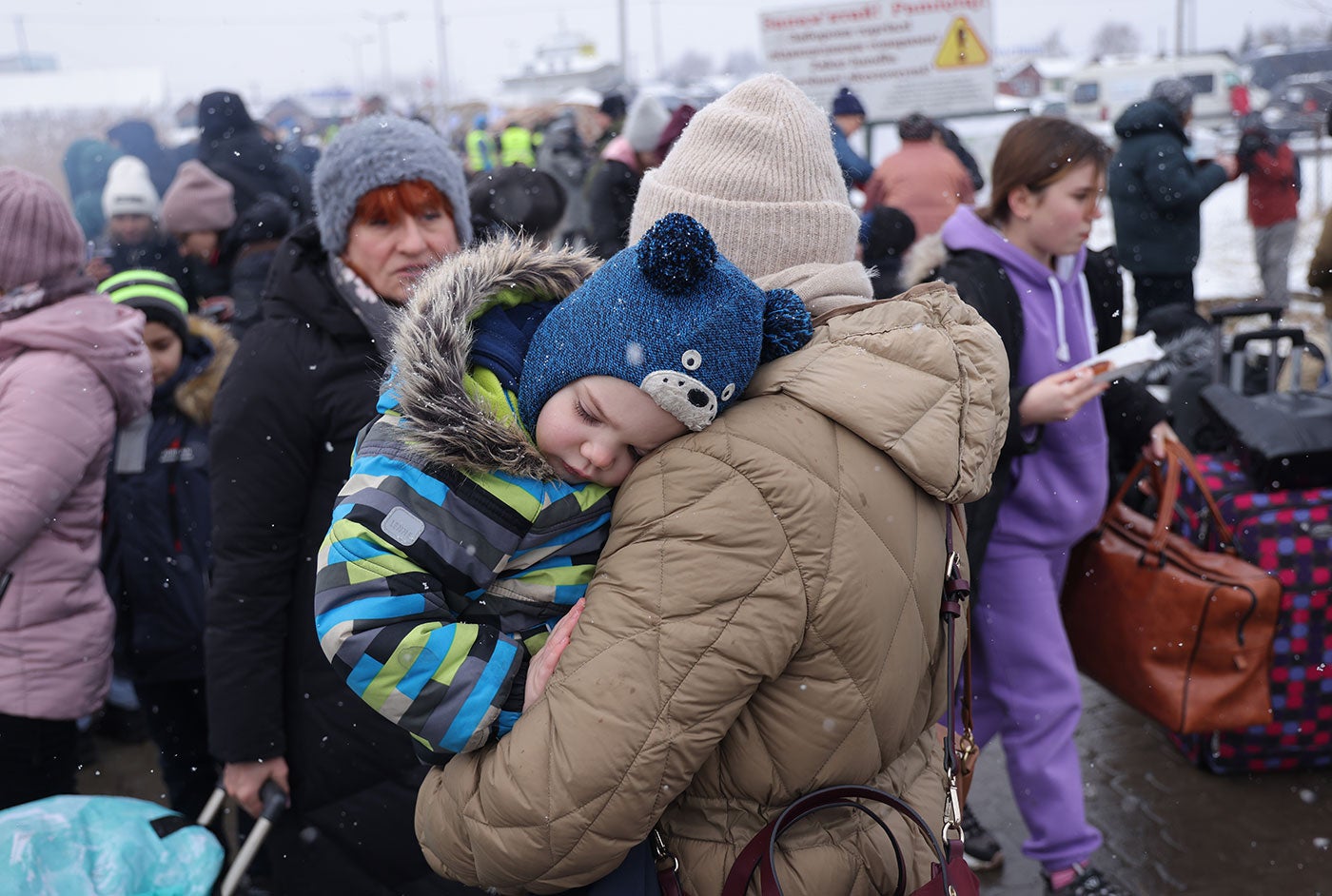 A large group of Ukrainians, many wearing winter clothing and carrying luggage, stand in a group. In the foreground, a parent holds a child.
