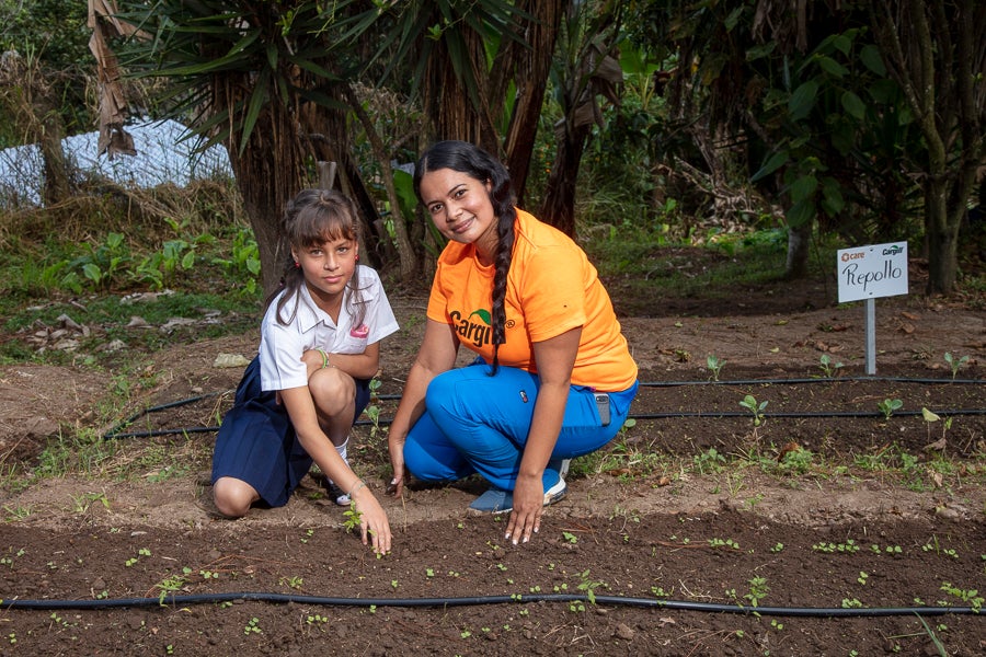 A woman and a girl squat near a row of seedlings.