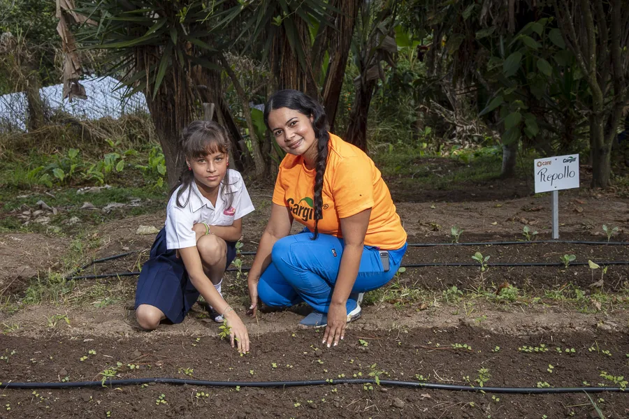 A woman and a girl squat near a row of seedlings.