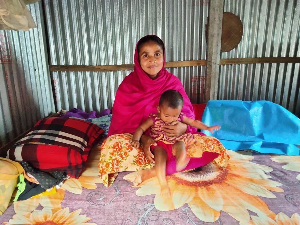 Una mujer de Bangladesh se sienta en una alfombra floral y sonríe mientras sostiene a su hijo pequeño.