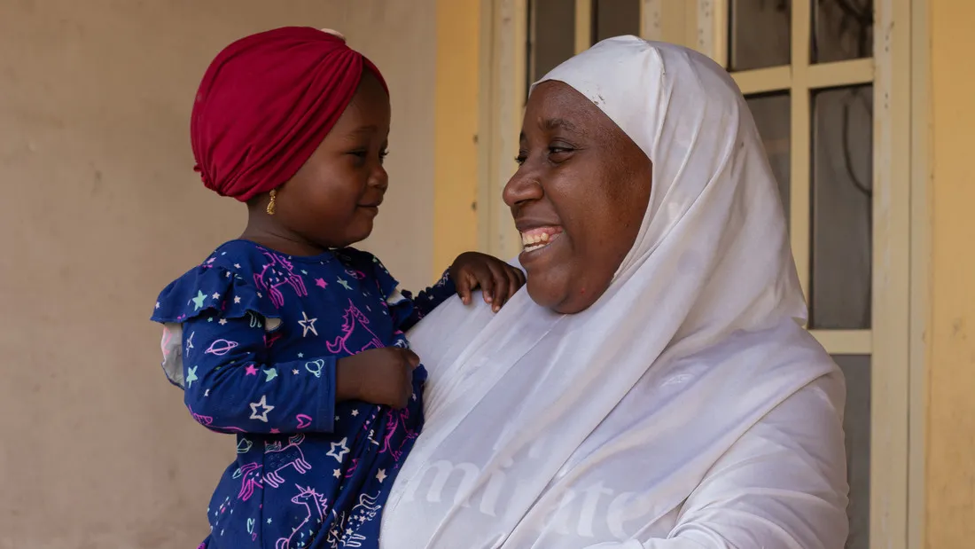 Uma mulher nigeriana toda vestida de branco sorri amplamente para sua filha, que ela está segurando em seus braços.