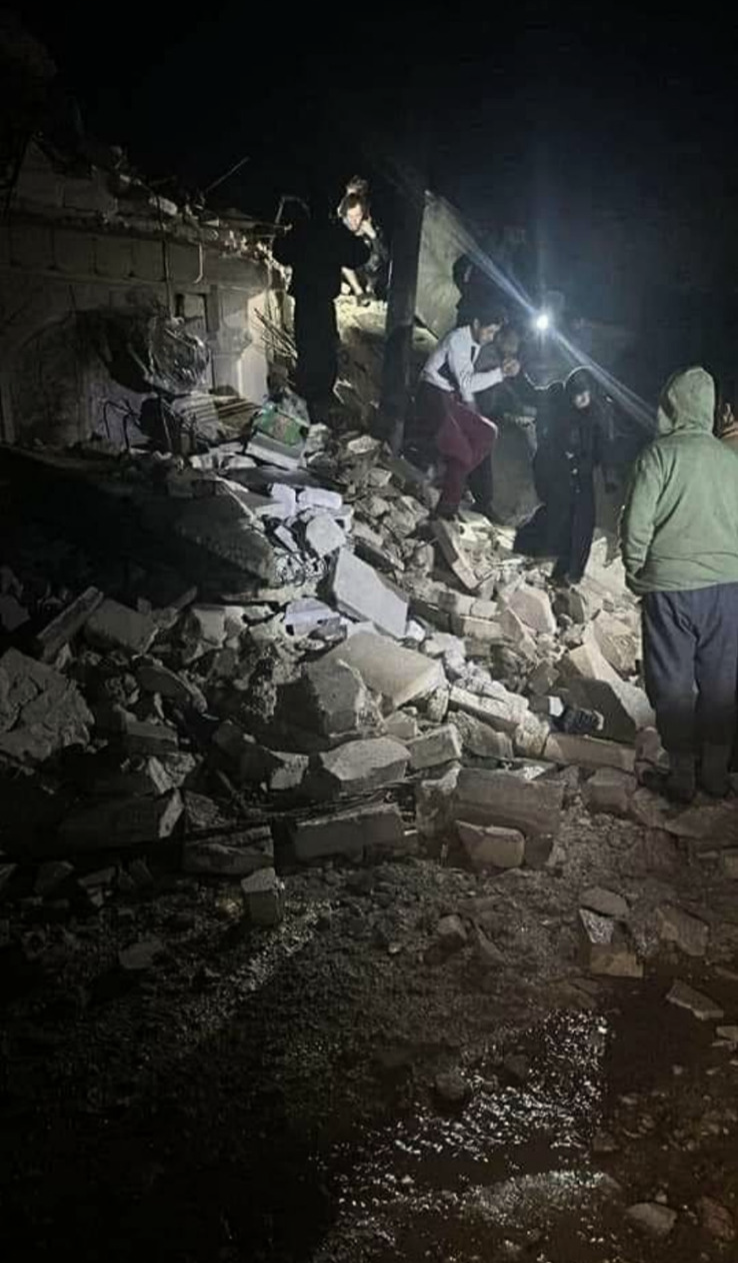 A group of people walk over building debris. It is dark outside, but there are bright lights shining on the people and rubble.