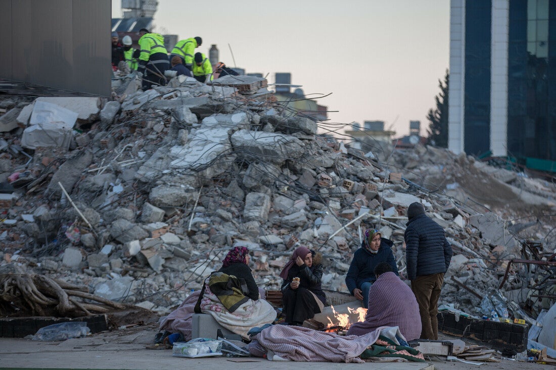 A Turkish family huddles under blankets by a small fire. Behind them is a large pile of rubble that emergency crew members wearing bright yellow vests are climbing over.