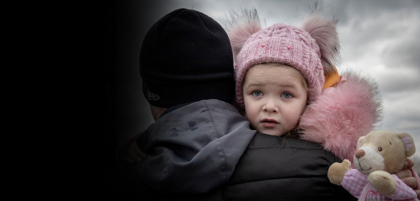 A young girl wearing a light pink winter coat and matching hat looks over the shoulder of the man carrying her.