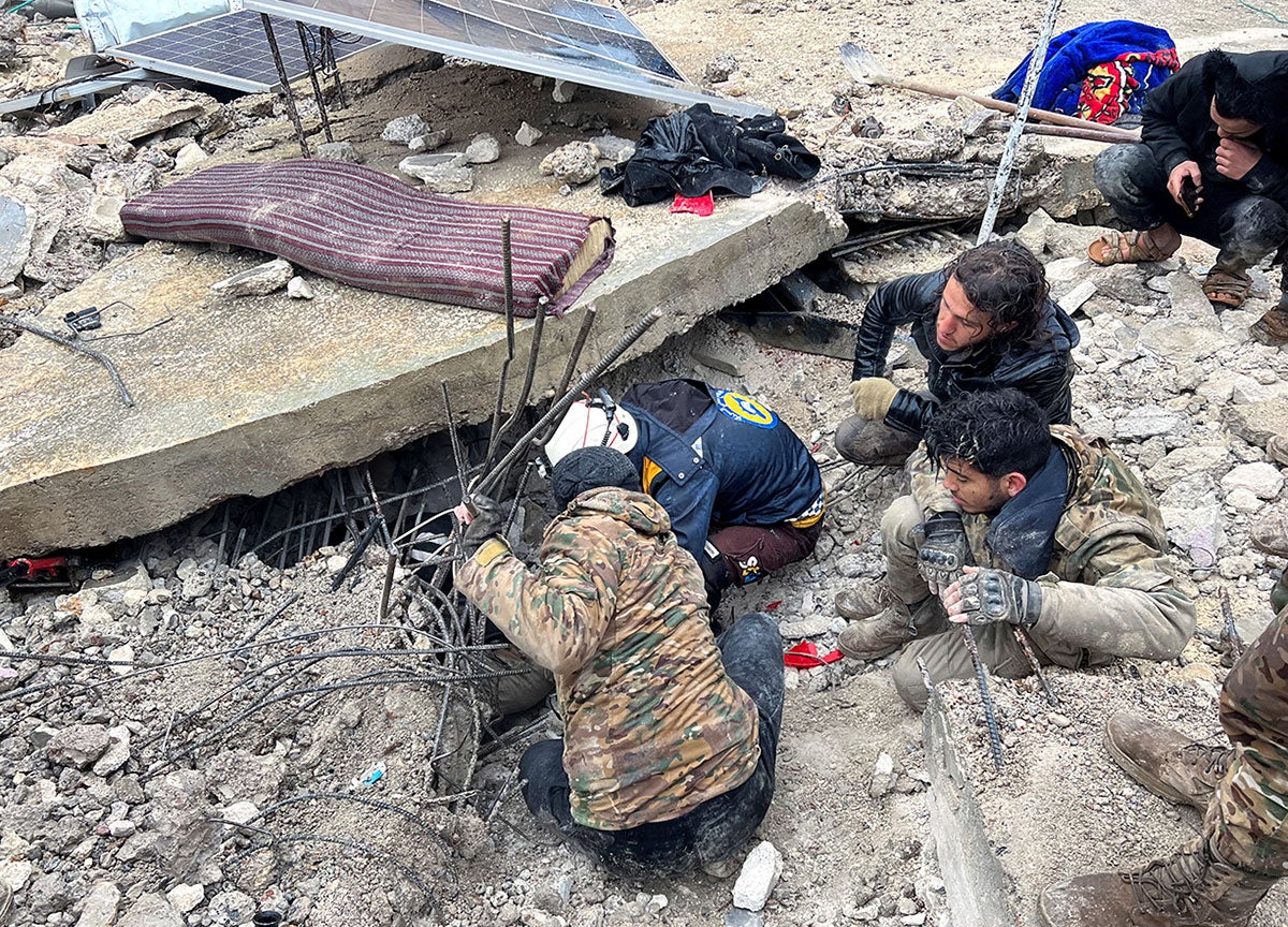 Un grupo de personas se arrodilla sobre los escombros y mira debajo de grandes pedazos de escombros de construcción.