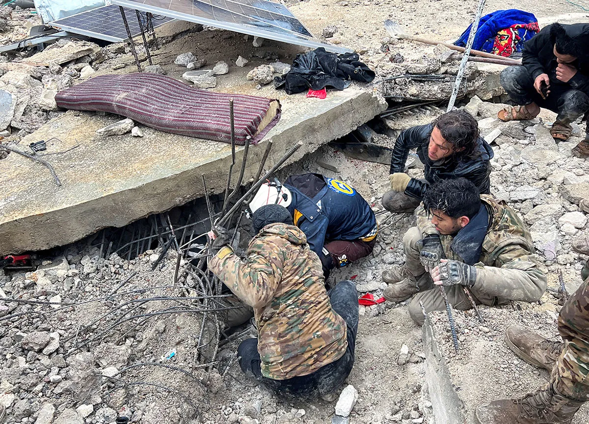 A group of people kneel on rubble and look underneath large pieces of building debris.