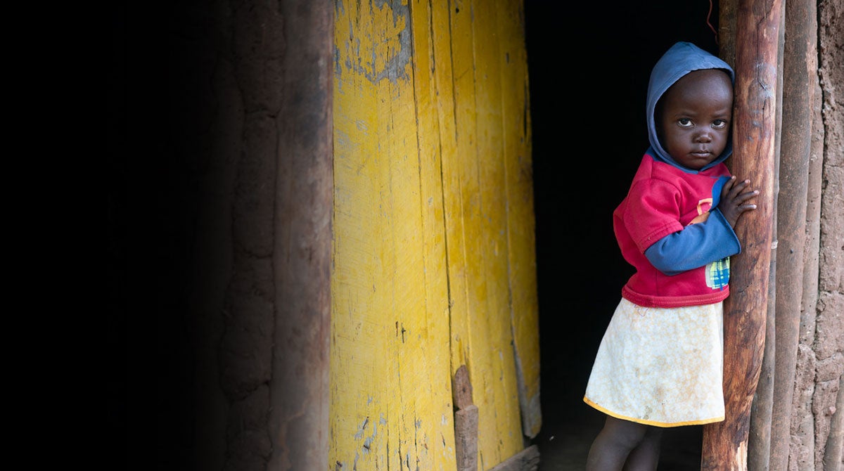 A young Zimbabwean girl peers around the corner of a doorway in a yellow wooden house.