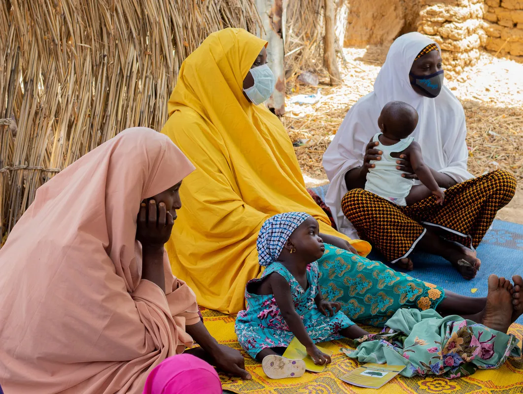 Three women sit on blankets on the ground with two small children.