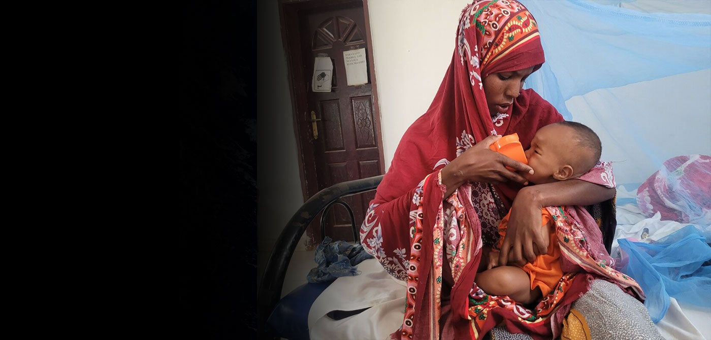 A Somalian woman wearing a red head scarf holds her baby and feeds him with an orange cup.