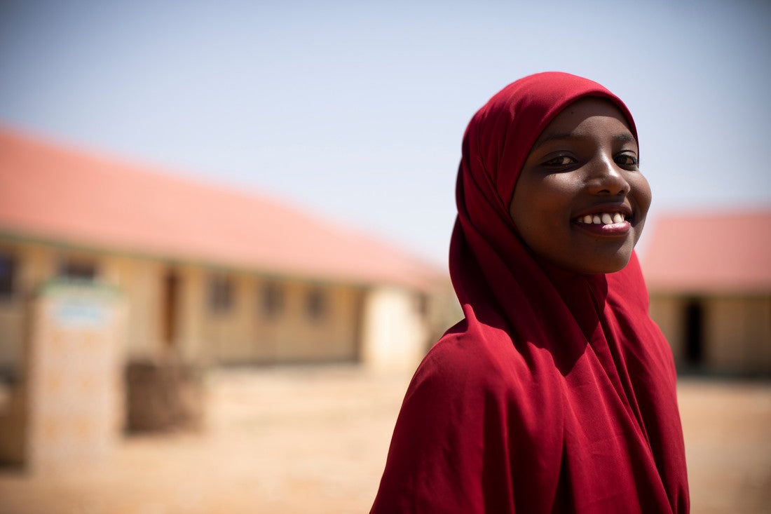 A girl wearing a dark red headscarf smiles at the camera. Behind her is a school.