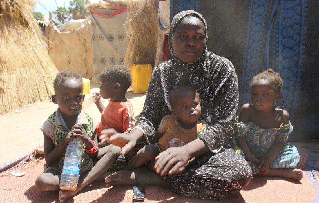 A woman sits on the ground with a small child sitting in her lap. Three other children sit around her, one of whom is holding a plastic water bottle.