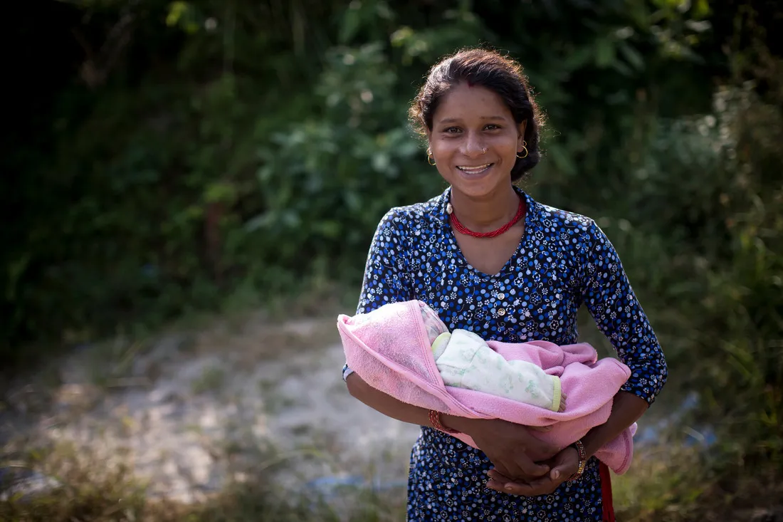 A Nepali woman wearing a blue dress smiles while holding a baby wrapped in a light pink blanket.