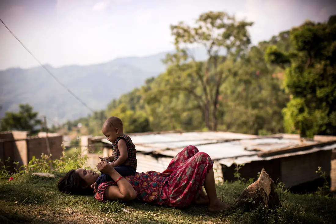 Une femme népalaise est allongée sur le dos dans un champ herbeux. Un bébé est assis sur sa poitrine et ils se tiennent la main.