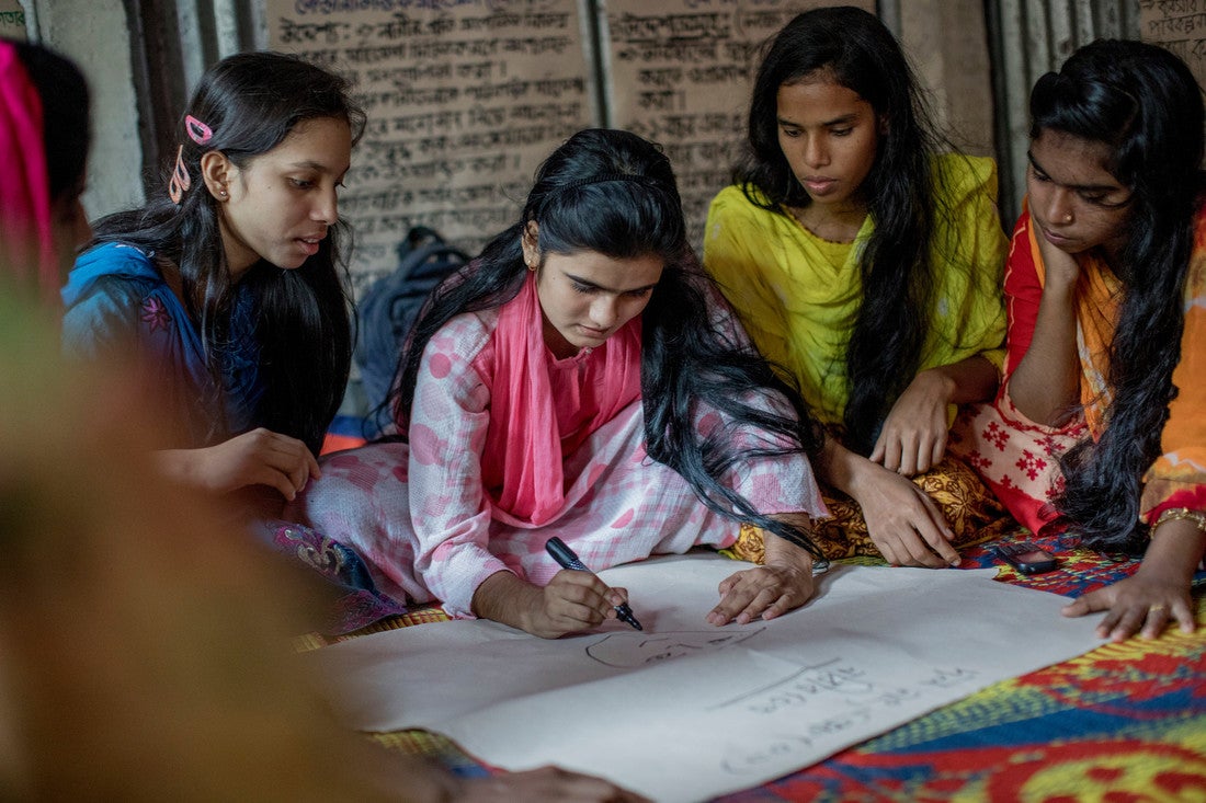 A group of Bangladeshi girls sit on the ground around a large piece of white paper. One girl, in the center, is drawing a face on the paper with a black marker.