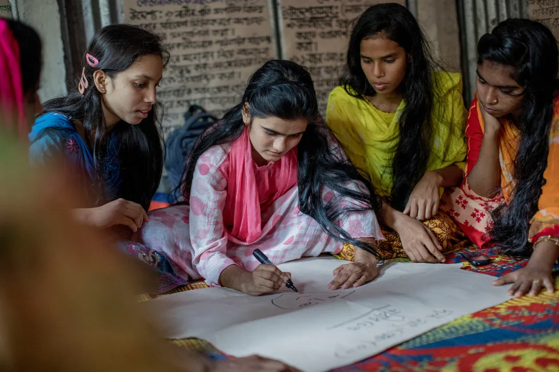 Um grupo de meninas de Bangladesh está sentado no chão em volta de um grande pedaço de papel branco. Uma garota, no centro, está desenhando um rosto no papel com um marcador preto.