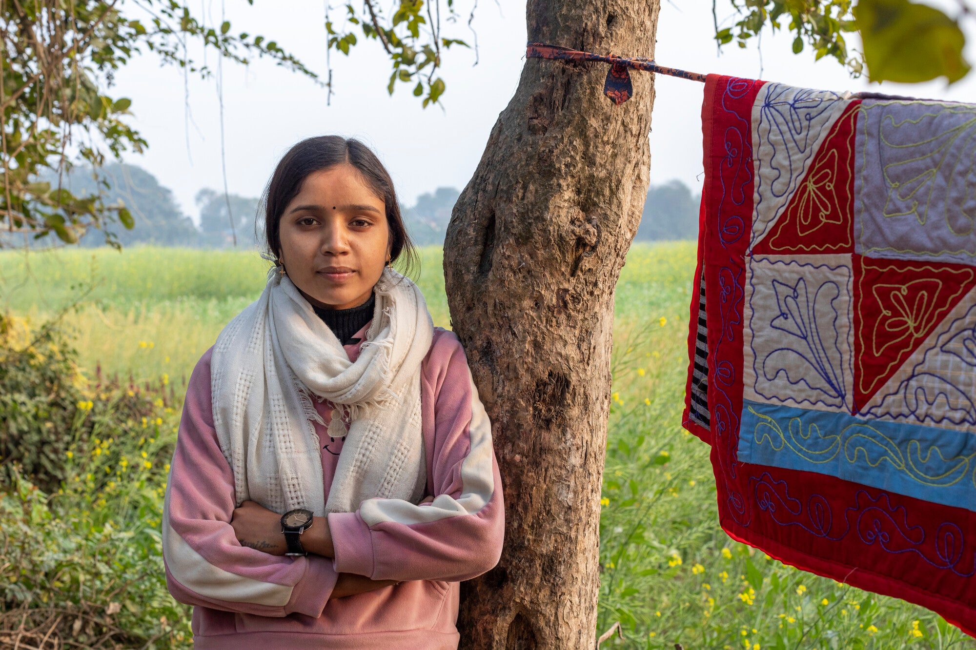 A young Nepali woman wearing a white scarf and pink jacket leans against a tree. A red and white quilt is hanging on a clothesline from the tree.