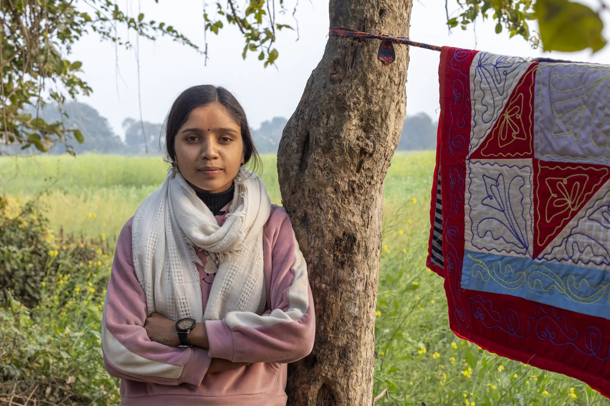 A young Nepali woman wearing a white scarf and pink jacket leans against a tree. A red and white quilt is hanging on a clothesline from the tree.