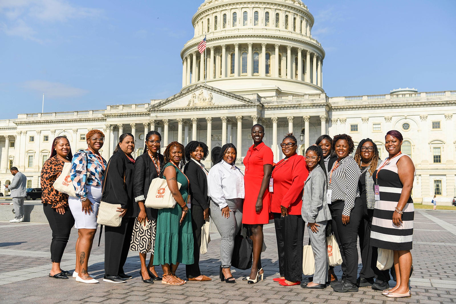 A group of Black women stand together and pose in front of the U.S. Capitol Building. A few are wearing CARE tote bags.