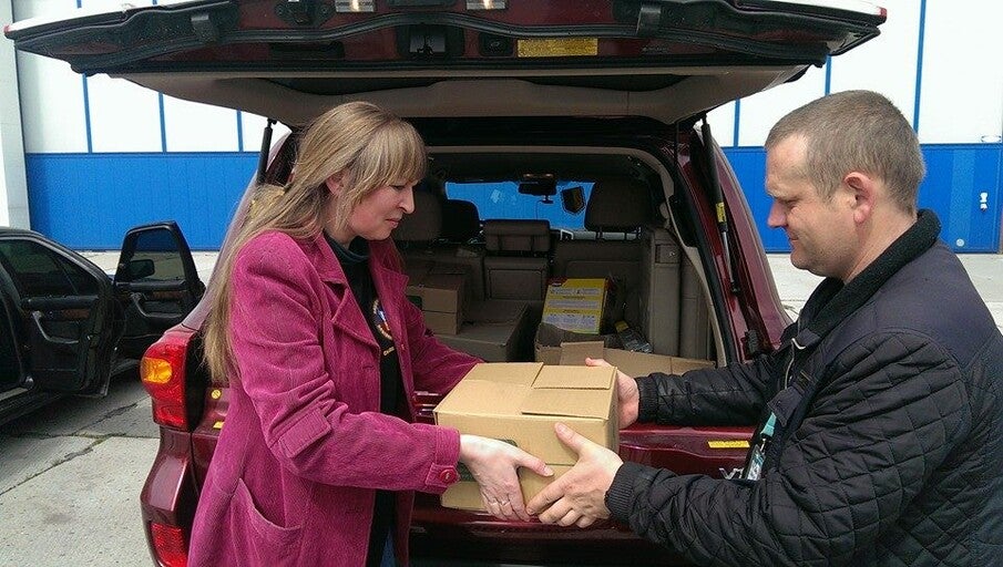 A woman hands a box to a man in front of a car.