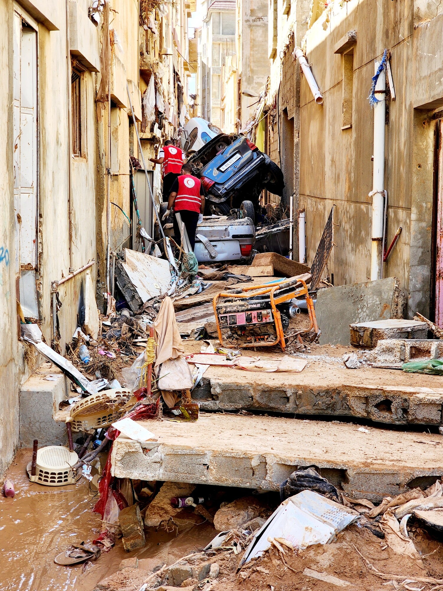 Narrow street between rows of buildings littered with debris, including two vehicles. Two men in red vests on the side with their backs to camera.