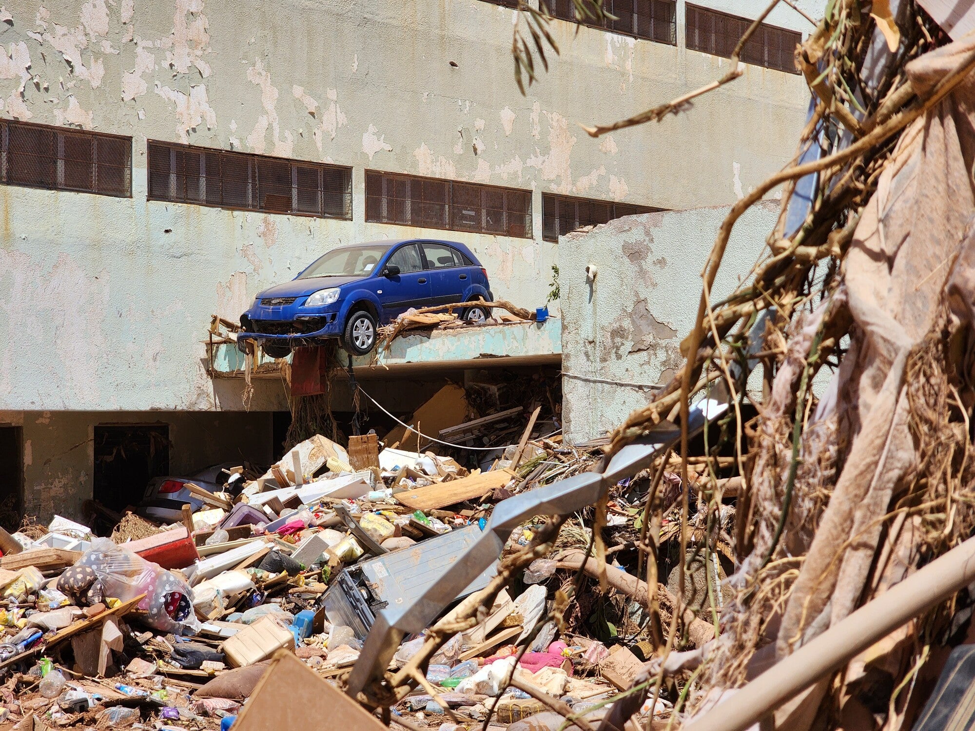 Rubble in foreground, building in background, blue car suspended on a ledge next to building.