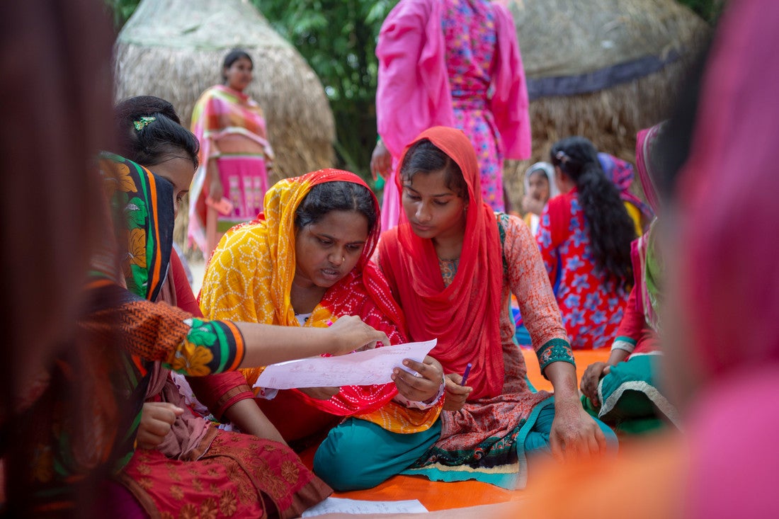 A group of young Bangladeshi women wearing colorful, body-length clothing sit together and review a document.
