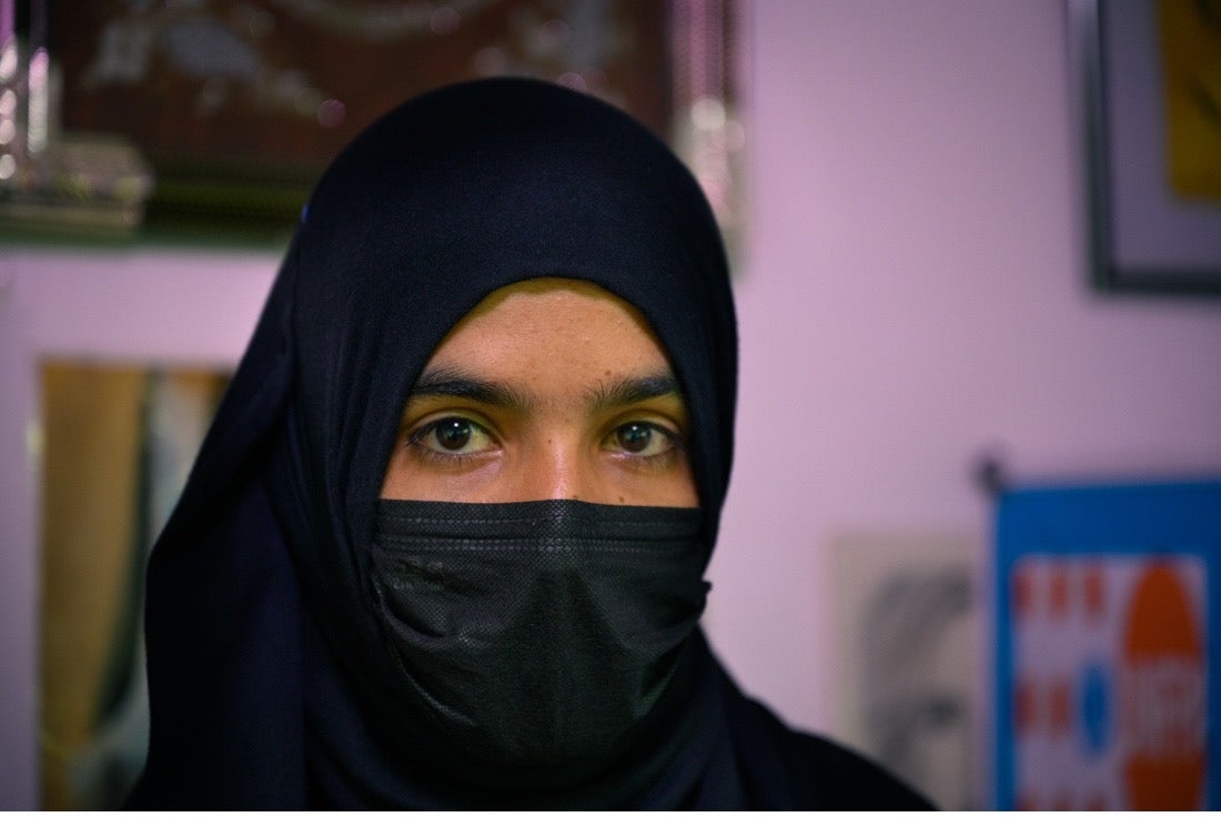 Close-up of young woman with head covering and mask, looking directly at the camera.
