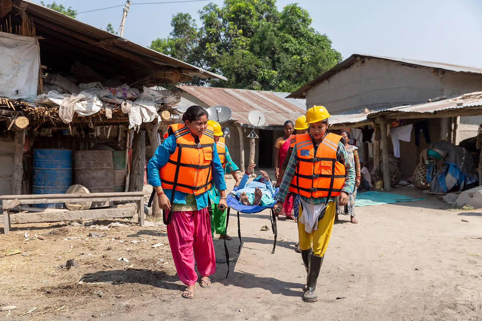 Two emergency workers wearing orange vests carry a stretcher through a village.