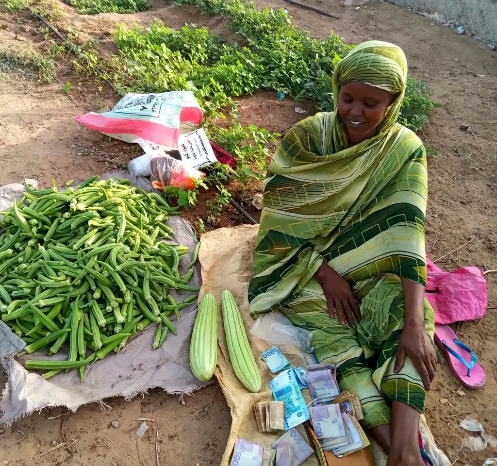 Una mujer vestida de verde y cubierta para la cabeza se sienta en el suelo entre verduras cosechadas