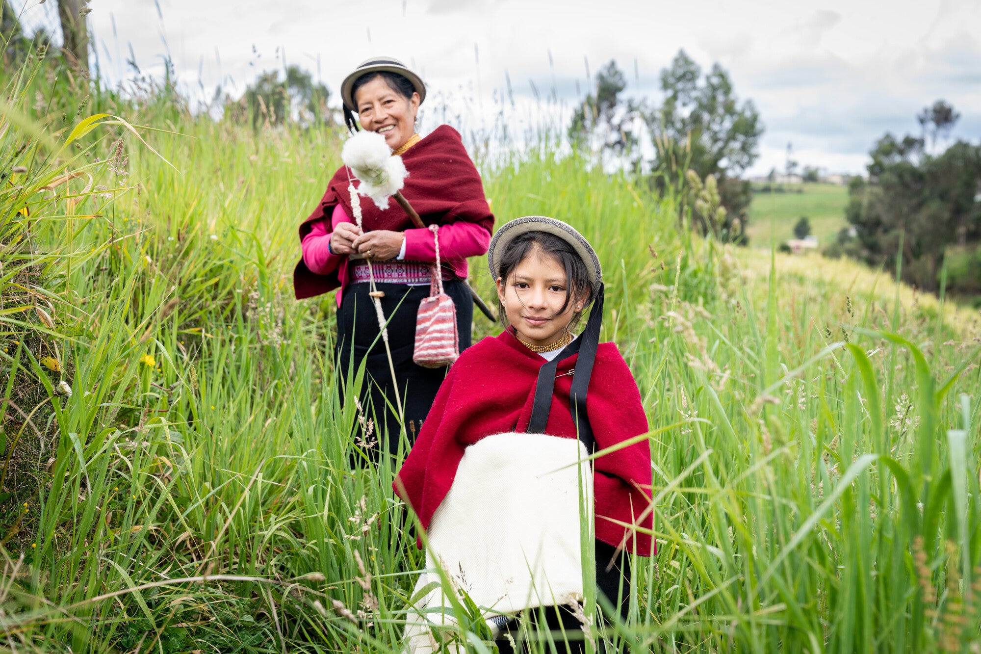 Woman and granddaughter in traditional Ecuadorian dress, standing on a grassy hillside.